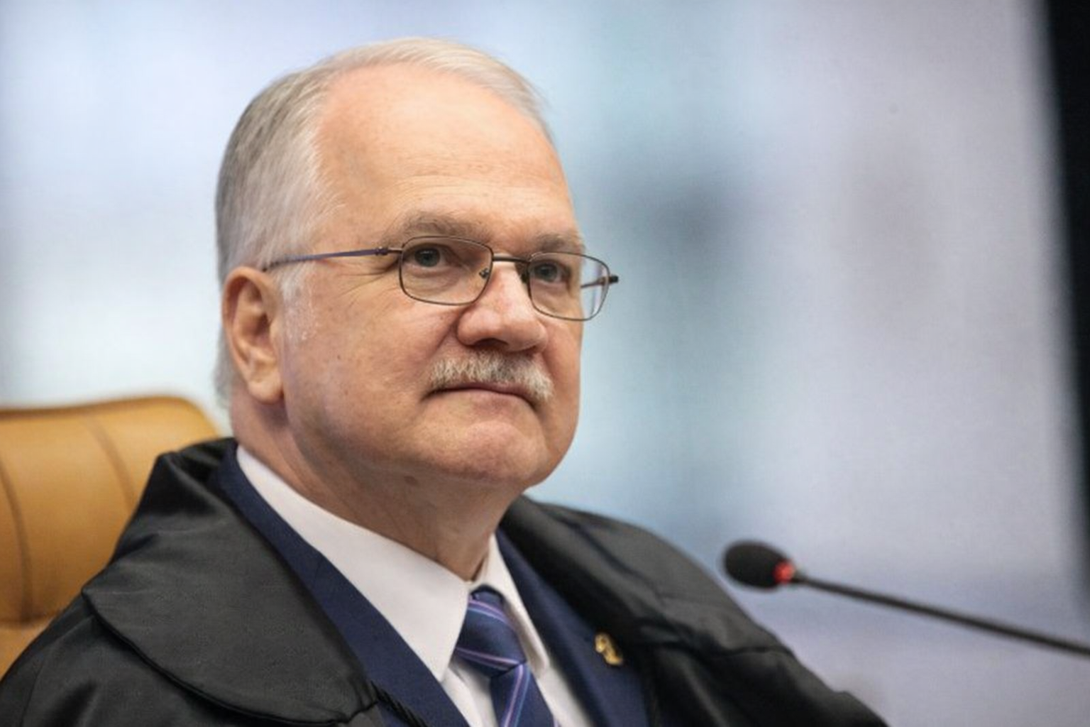 Retrato em três quartos do ministro Edson Fachin no plenário do STF, usando toga, gravata azul e sentado em cadeira marrom com microfone ao lado.