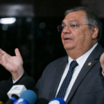 Flávio Dino em coletiva de imprensa, gesticulando com as mãos abertas diante de microfones, em enquadramento lateral e fundo escuro.