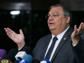 Flávio Dino em coletiva de imprensa, gesticulando com as mãos abertas diante de microfones, em enquadramento lateral e fundo escuro.