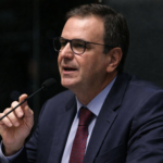 Eduardo Paes falando ao microfone em sessão oficial, usando terno escuro e gravata vermelha, em enquadramento lateral com fundo institucional.