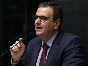 Eduardo Paes falando ao microfone em sessão oficial, usando terno escuro e gravata vermelha, em enquadramento lateral com fundo institucional.