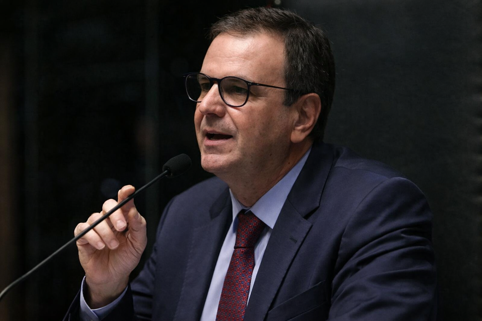 Eduardo Paes falando ao microfone em sessão oficial, usando terno escuro e gravata vermelha, em enquadramento lateral com fundo institucional.