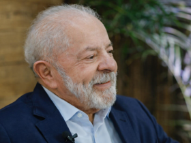 Luiz Inácio Lula da Silva em retrato lateral, sorrindo levemente, usando terno azul e camisa clara, com fundo interno desfocado e plantas ao fundo.