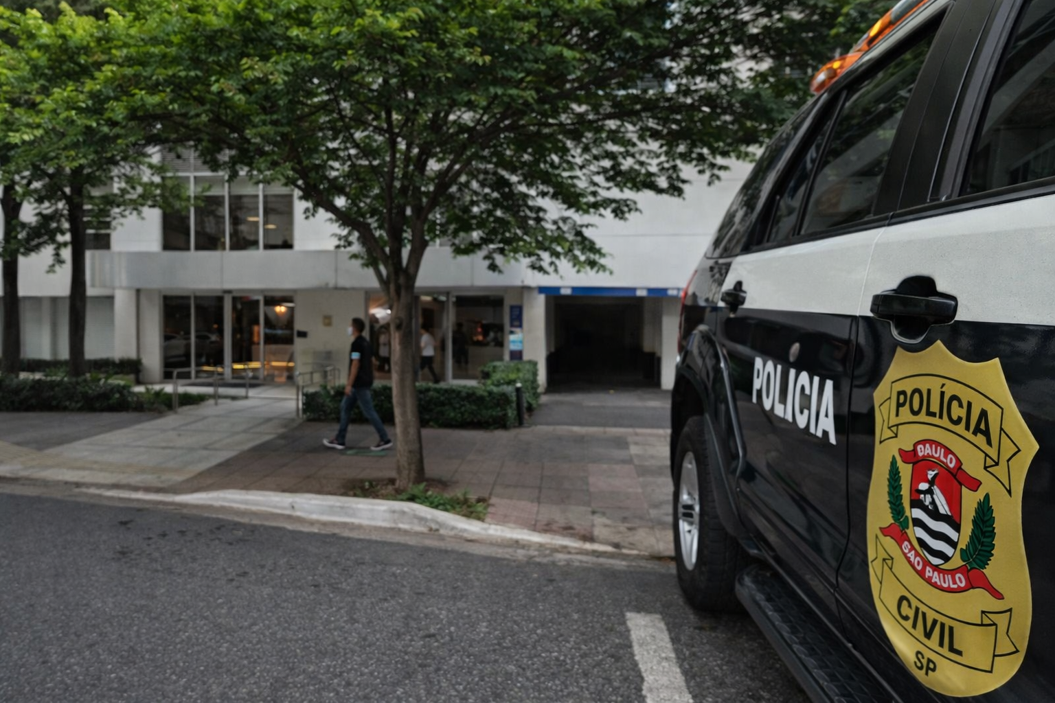 Entrada de prédio comercial em Moema, São Paulo, com fachada branca, árvore em frente e pedestre caminhando na calçada.