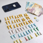 Entorpecentes embalados em pinos coloridos, dinheiro em reais e celular apreendidos durante ação policial, organizados sobre mesa branca.