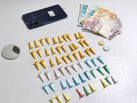 Entorpecentes embalados em pinos coloridos, dinheiro em reais e celular apreendidos durante ação policial, organizados sobre mesa branca.