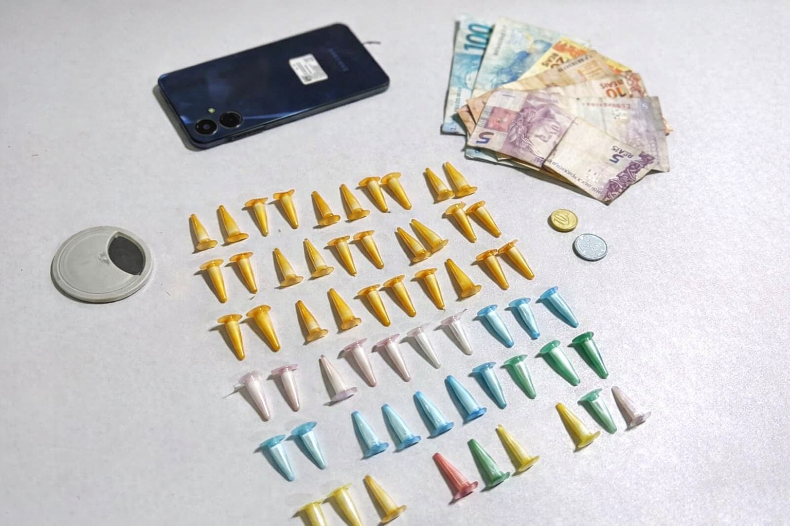 Entorpecentes embalados em pinos coloridos, dinheiro em reais e celular apreendidos durante ação policial, organizados sobre mesa branca.