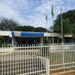 Fachada de unidade da Polícia Militar em Curvelo, Minas Gerais, com bandeiras hasteadas e grade branca na parte frontal do prédio.