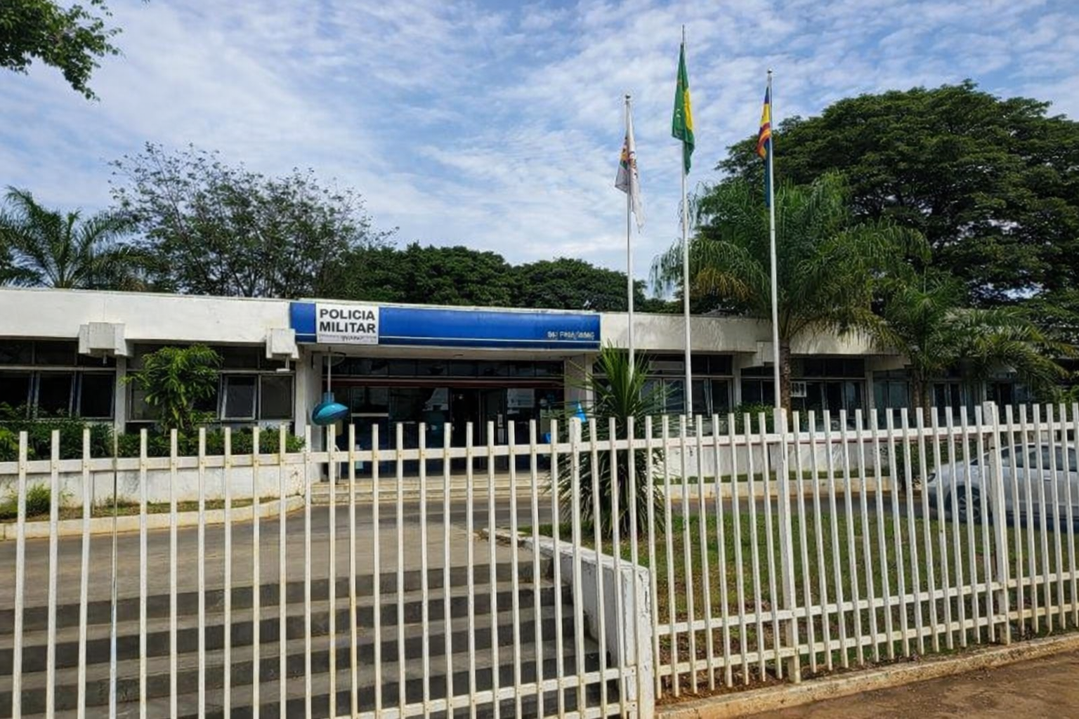 Fachada de unidade da Polícia Militar em Curvelo, Minas Gerais, com bandeiras hasteadas e grade branca na parte frontal do prédio.