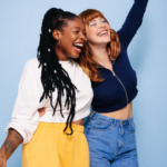 Duas mulheres jovens sorrindo e abraçadas diante de fundo azul, representando amizade, diversidade e alegria