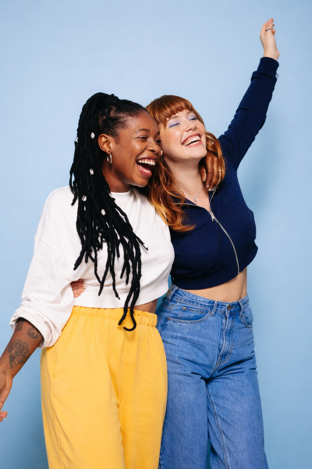Duas mulheres jovens sorrindo e abraçadas diante de fundo azul, representando amizade, diversidade e alegria