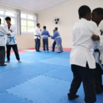 Estudantes treinando judô em sala com tatame durante projeto de contraturno escolar em ambiente educativo.