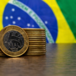 Pilha de moedas de 1 real sobre mesa com bandeira do Brasil ao fundo, simbolizando economia e balança comercial brasileira.