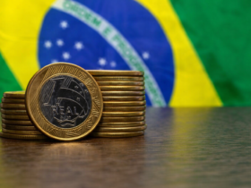 Pilha de moedas de 1 real sobre mesa com bandeira do Brasil ao fundo, simbolizando economia e balança comercial brasileira.