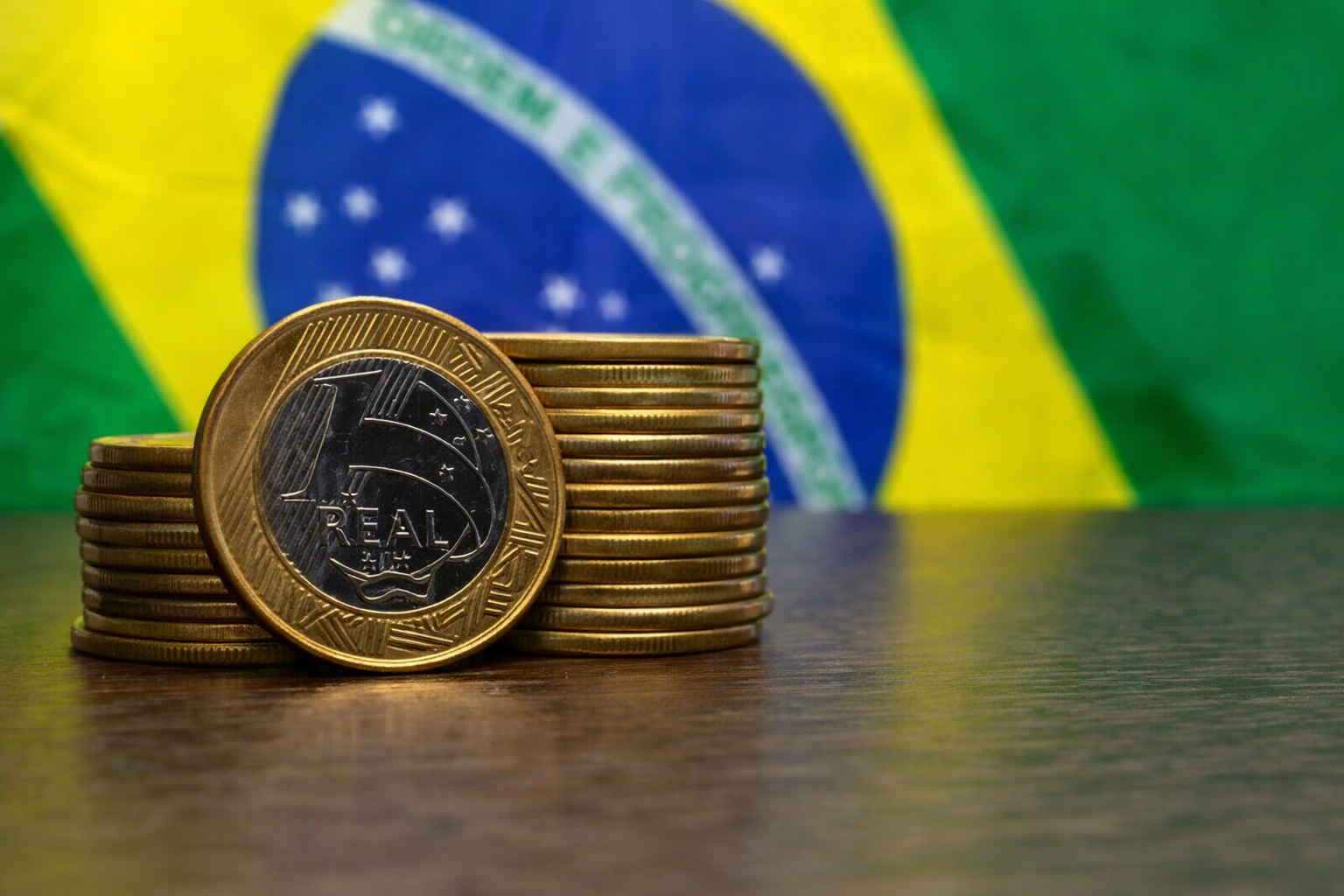 Pilha de moedas de 1 real sobre mesa com bandeira do Brasil ao fundo, simbolizando economia e balança comercial brasileira.