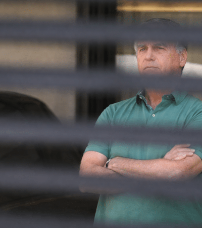 Bolsonaro visto através de persiana em imagem que simboliza acompanhamento médico e momento de incerteza política no Brasil